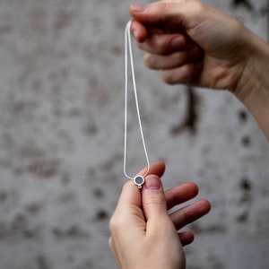 Puede incluir: Un collar de cadena de plata con un peque&ntilde;o colgante redondo. El colgante es negro y tiene un anillo de plata alrededor.