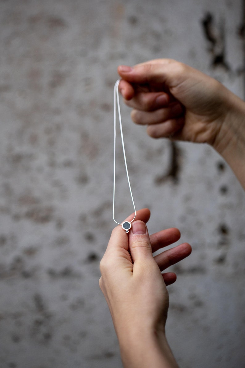 Puede incluir: Un collar de cadena de plata con un peque&ntilde;o colgante redondo. El colgante es negro y tiene un anillo de plata alrededor.