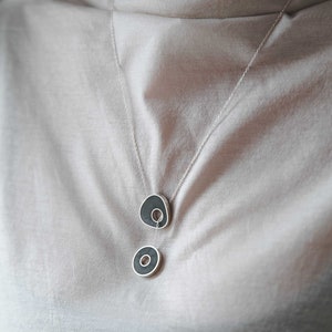 May include: A silver chain necklace with two black ceramic pendants. The top pendant is an irregular shape with a hole in the center. The bottom pendant is a circle with a hole in the center.