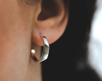 May include: Silver hoop earrings with a geometric, faceted design. The earrings are worn in a person's ear.