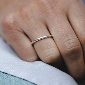 May include: A close-up of a silver ring on a finger. The ring is a thin band with a smooth, polished surface. The ring is worn on a finger, with a white fabric and denim background.