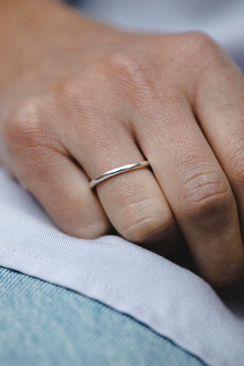 May include: A close-up of a silver ring on a finger. The ring is a thin band with a smooth, polished surface. The ring is worn on a finger, with a white fabric and denim background.