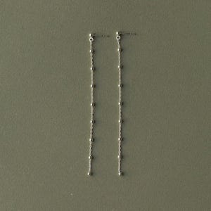 Puede incluir: Un par de pendientes colgantes de tono plateado con un diseño de cadena larga y delicada. Cada pendiente consta de una fina cadena vertical adornada con pequeñas cuentas esféricas espaciadas uniformemente y un cierre de tipo poste.