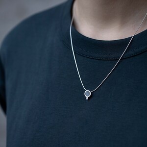 Puede incluir: Un collar de cadena de plata con un peque&ntilde;o colgante redondo. El colgante es negro con un c&iacute;rculo plateado en el centro.