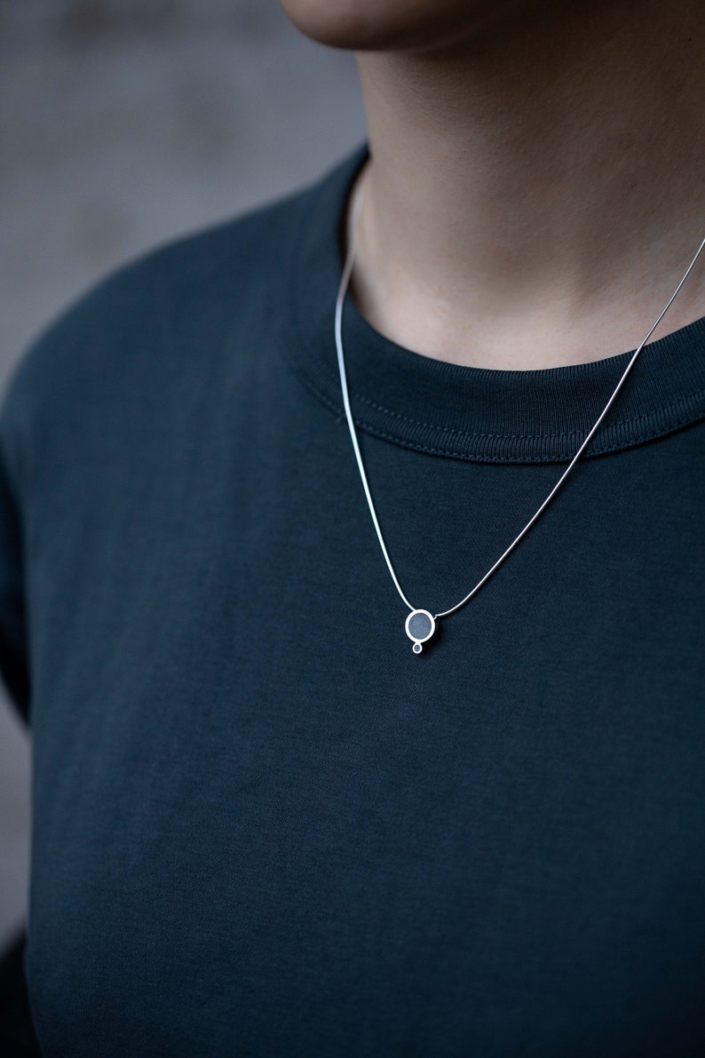 Puede incluir: Un collar de cadena de plata con un peque&ntilde;o colgante redondo. El colgante es negro con un c&iacute;rculo plateado en el centro.