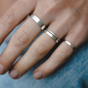 Puede incluir: Una mano con cuatro anillos de plata. Los anillos están apilados en dos dedos. Los anillos son simples y minimalistas.