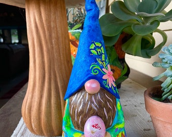 May include: A blue and green gnome with a long blue hat holds a pink Easter egg. The gnome has a brown beard and a pink nose. The gnome is standing in front of a red and white mushroom.