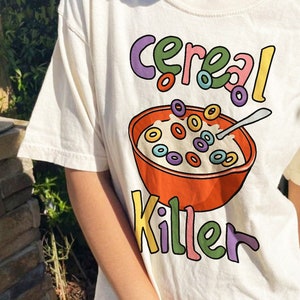 May include: Off-white t-shirt featuring a colorful graphic of a bowl of cereal with the words "Cereal Killer" in playful lettering. The cereal is in a red bowl with a spoon.