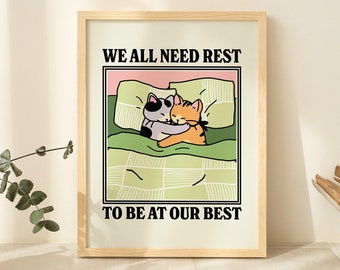 May include: A framed print with a light background and black text that reads "We all need rest to be at our best". The print features a cartoon illustration of two cats cuddling in bed.