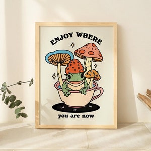 May include: A framed print with a colorful illustration of a frog sitting in a teacup surrounded by mushrooms. The text "Enjoy where you are now" is printed below the illustration.