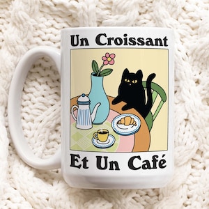 Könnte beinhalten: Weiße Keramiktasse mit einer Illustration einer schwarzen Katze. Die Katze sitzt auf einem Stuhl an einem Tisch mit einem Croissant, einer Kaffeetasse und einer Teekanne. Der Text auf der Tasse lautet "Un Croissant Et Un Café".