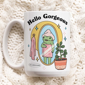 May include: White ceramic mug featuring a frog in a towel looking in a mirror. The mug has the text "Hello Gorgeous" and "You Look So Good" on it.