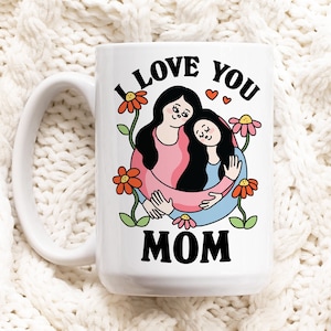 K&ouml;nnte beinhalten: Eine wei&szlig;e Keramiktasse mit einer Schwarzwei&szlig;illustration von zwei Frauen, die sich umarmen. Der Text "I LOVE YOU MOM" ist in Schwarz auf die Tasse gedruckt.