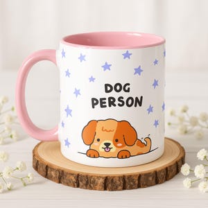 Op de afbeelding: Een witte keramische mok met een roze handvat en binnenkant. De mok heeft een cartoonhondenillustratie en de tekst "DOG PERSON" met blauwe steraccenten. De mok staat op een houten onderzetter.