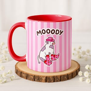 May include: A ceramic mug with a red handle and interior. The exterior features pink and white vertical stripes and a cartoon cow wearing a pink hat and boots. The word "MOOODY" is printed above the cow.