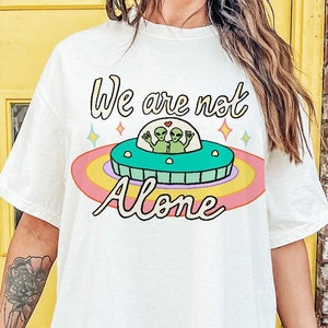 May include: White oversized t-shirt with a graphic of two green aliens in a flying saucer with the text "We are not alone" in pink and blue lettering.