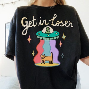 May include: Black t-shirt with a graphic of a UFO with a green alien inside, a cat standing on a rainbow beam, and the text "Get in Loser".