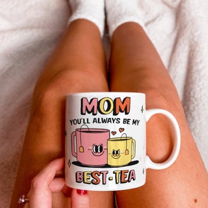 May include: White ceramic mug with a pink and yellow teacup design. The mug reads "Mom you'll always be my best-tea".