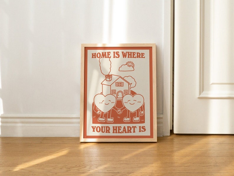 May include: A framed print with a white background and red text that reads "Home is where your heart is". The print features a cartoon illustration of two hearts holding hands in front of a house with smoke coming from the chimney.