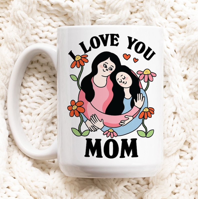 K&ouml;nnte beinhalten: Eine wei&szlig;e Keramiktasse mit einer Schwarzwei&szlig;illustration von zwei Frauen, die sich umarmen. Der Text "I LOVE YOU MOM" ist in Schwarz auf die Tasse gedruckt.