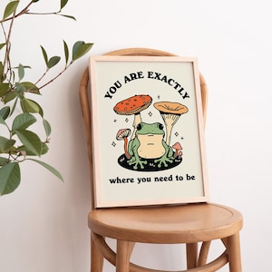 May include: A framed print featuring a green frog perched on a mushroom. The print includes the text "You are exactly where you need to be" in black lettering.