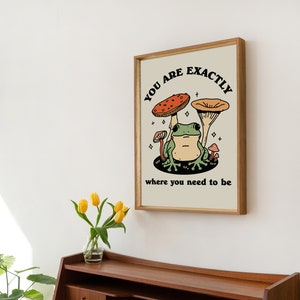 May include: A framed print featuring a green frog sitting beneath two mushrooms. The text on the print reads "You are exactly where you need to be".