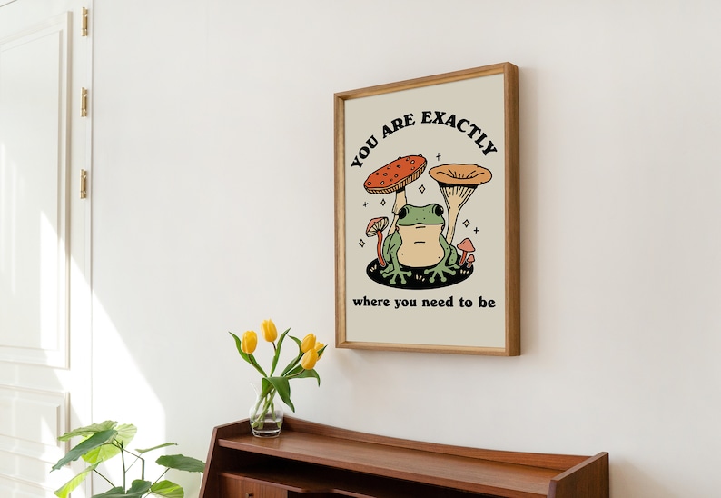 May include: A framed print featuring a green frog sitting beneath two mushrooms. The text on the print reads "You are exactly where you need to be".
