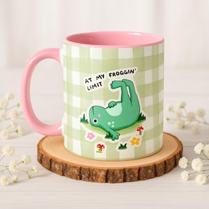 May include: A ceramic mug with a pink handle and interior. The exterior features a green and white checkered pattern with a cartoon frog illustration and the text "AT MY FROGGIN' LIMIT". The mug is placed on a wooden coaster.
