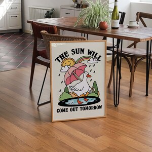 Op de afbeelding: Een ingelijste illustratie met een witte gans die een roze paraplu vasthoudt. De illustratie is geplaatst tegen een achtergrond van een regenboog, wolken en regendruppels. De tekst "THE SUN WILL COME OUT TOMORROW" is onder de afbeelding gedrukt.