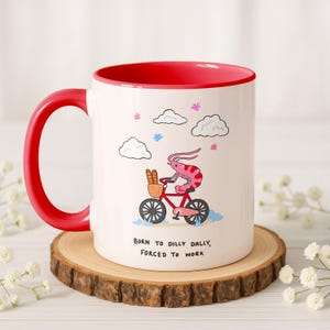 Op de afbeelding: Witte keramische mok met een rode binnenkant en handvat. De mok heeft een grillige illustratie van een garnaal op een rode fiets met een broodmand, wolken en de tekst "BORN TO DILLY DALLY, FORCED TO WORK".