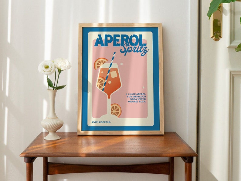 May include: A framed print with a pink background and blue and white text that reads "Aperol Spritz". The print features an illustration of a glass of Aperol Spritz with orange slices and a straw. The text below the illustration reads "1 1/4 oz Aperol, 2 oz Prosecco, Soda Water, Orange Slice" and "Cyn Cocktail".