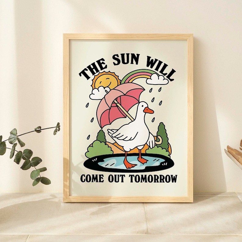 Op de afbeelding: Een ingelijst print met een witte achtergrond en een cartoonillustratie van een gans die een paraplu vasthoudt. De tekst "THE SUN WILL COME OUT TOMORROW" is onder de gans afgedrukt.