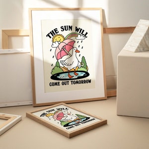 Op de afbeelding: Een ingelijste illustratie van een witte gans met een roze paraplu die in een plas water staat. De tekst "The sun will come out tomorrow" staat onder de gans.