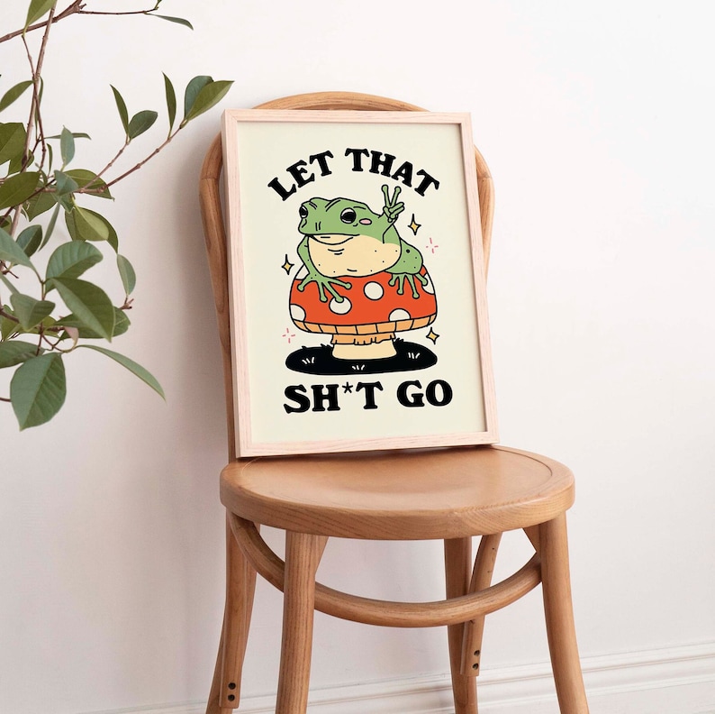 Pu&ograve; includere: Una stampa incorniciata con uno sfondo giallo chiaro che presenta una rana di cartone animato seduta su un fungo rosso e bianco. La rana sta facendo il segno della pace e il testo "Let That Sh*t Go" &egrave; scritto in nero sotto il fungo.