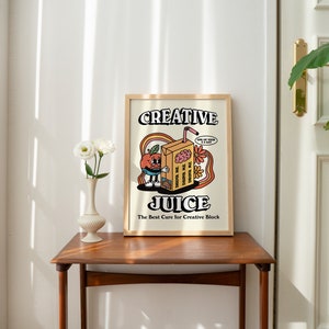 May include: A framed print with a cartoon illustration of a juice box with a face and the text "Creative Juice. The Best Cure for Creative Block." The juice box is holding a brain and has a thought bubble that says "I'm making the world a better place, one sip at a time."