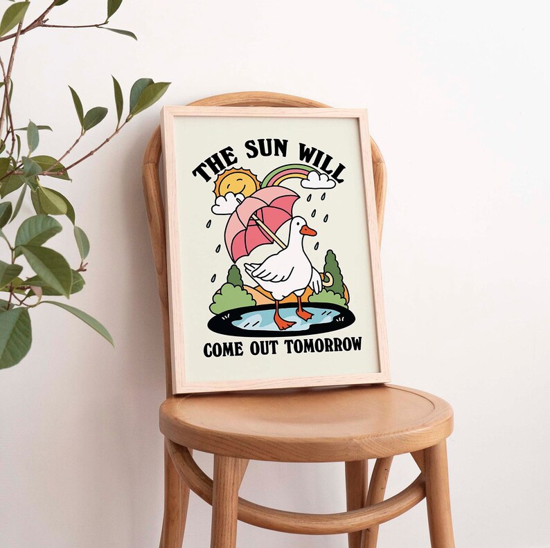 Op de afbeelding: Een ingelijst print met een cartoonillustratie van een witte eend die een paraplu vasthoudt. De tekst luidt "De zon komt morgen weer terug".