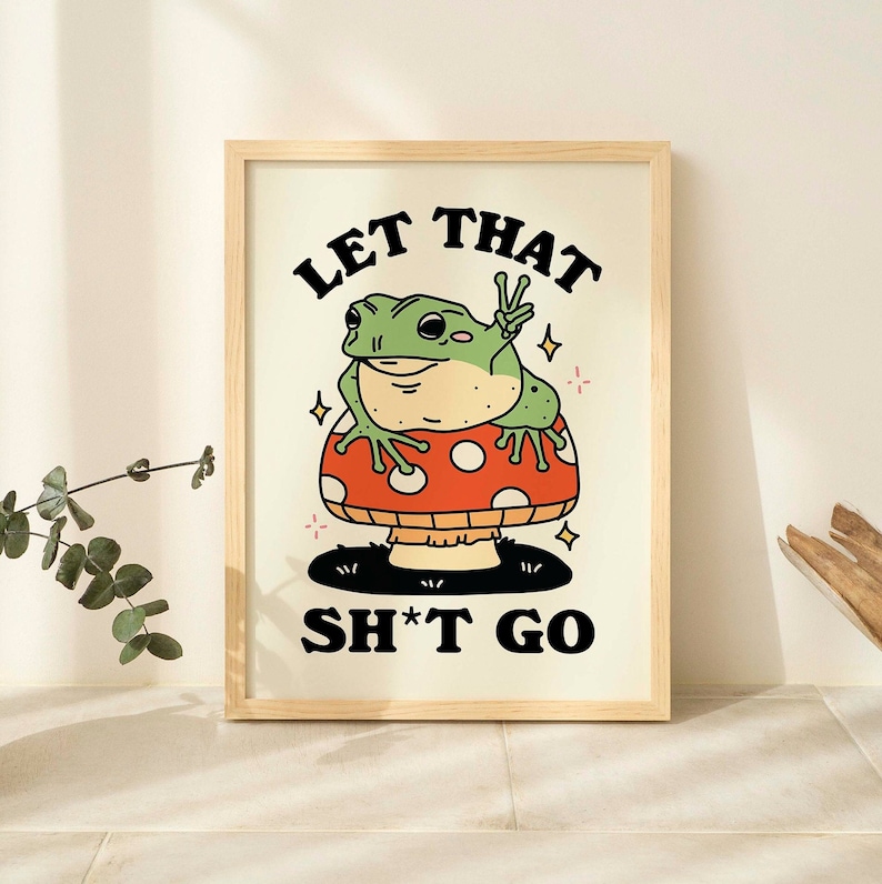 Pu&ograve; includere: Una stampa incorniciata con una rana verde seduta su un fungo rosso e bianco. Il testo "Let That Sh*t Go" &egrave; stampato in nero sotto la rana.