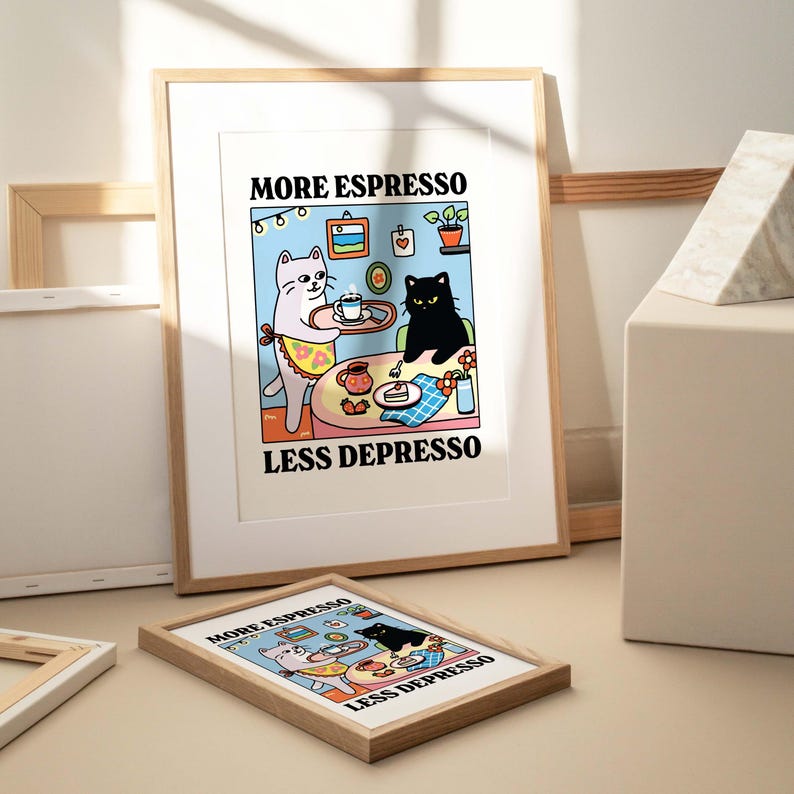 May include: A framed print with a cartoon illustration of two cats. The white cat is holding a tray with a cup of coffee. The black cat is sitting at a table with a slice of cake. The text on the print reads "More Espresso Less Depresso".