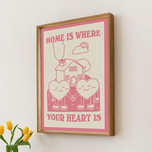 May include: A pink and white framed print with the text "Home is where your heart is". The print features a cartoon illustration of two hearts holding hands in front of a house with a chimney and smoke coming out of it. There is a sun and a cloud above the house.