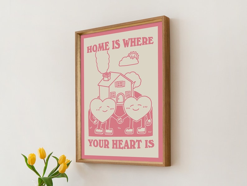 May include: A pink and white framed print with the text "Home is where your heart is". The print features a cartoon illustration of two hearts holding hands in front of a house with a chimney and smoke coming out of it. There is a sun and a cloud above the house.