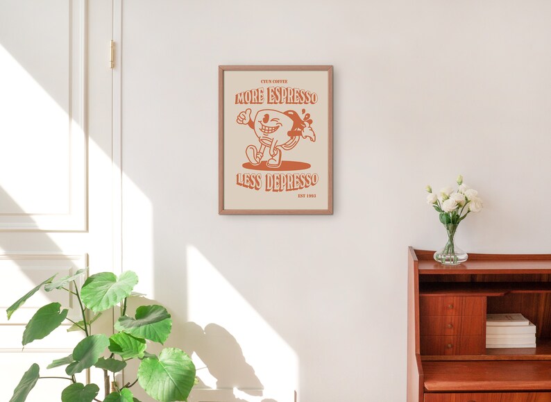 K&ouml;nnte beinhalten: Ein gerahmter Druck mit einer Cartoon-Illustration einer Kaffeetasse mit einem Gesicht und dem Text "More Espresso Less Depresso Est. 2003". Der Druck ist auf wei&szlig;em Hintergrund mit braunem Rahmen.