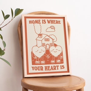 May include: A framed print with a beige background and orange text that reads "Home is where your heart is". The print features a cartoon illustration of a house with two hearts holding hands in front of it.