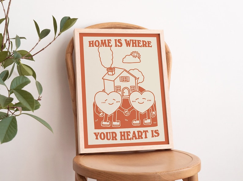 May include: A framed print with a beige background and orange text that reads "Home is where your heart is". The print features a cartoon illustration of a house with two hearts holding hands in front of it.
