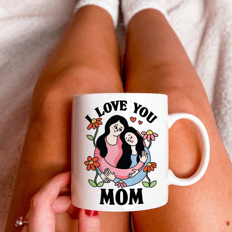 K&ouml;nnte beinhalten: Eine wei&szlig;e Keramiktasse mit einer schwarzen und roten Illustration von zwei Frauen, die sich umarmen. Der Text "I Love You Mom" ist auf die Tasse gedruckt.