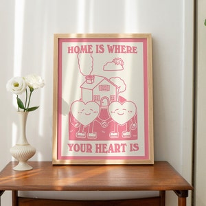 May include: Pink framed poster with a white background and the text "Home is where your heart is". The poster features a cartoon illustration of two hearts holding hands in front of a house with a chimney.