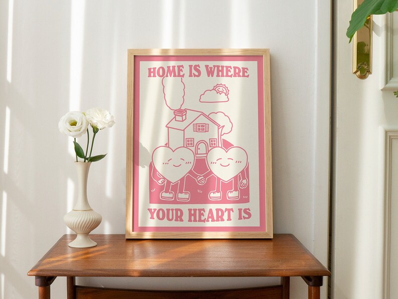 May include: Pink framed poster with a white background and the text "Home is where your heart is". The poster features a cartoon illustration of two hearts holding hands in front of a house with a chimney.