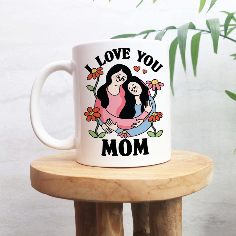 K&ouml;nnte beinhalten: Wei&szlig;e Keramiktasse mit einer Schwarzwei&szlig;illustration von zwei Personen, die sich umarmen. Der Text "I LOVE YOU MOM" ist in Schwarz auf die Tasse gedruckt.