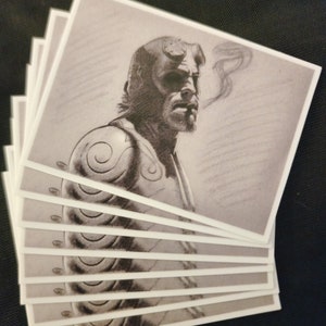 May include: A stack of eight black and white printed postcards featuring a close-up portrait of a man with horns and a cigarette in his mouth. The man has a serious expression and is wearing a detailed, patterned shirt.