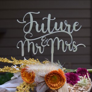 May include: Silver cake topper with the words "Future Mr & Mrs" in a cursive font. The topper is placed on a bouquet of dried flowers with orange and purple blooms.