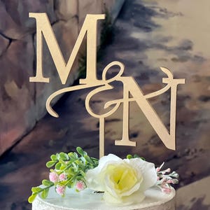 May include: A wooden cake topper featuring the intertwined letters "M" and "N". The letters are in a serif font and are light brown. The topper is decorated with white and yellow flowers, and greenery. A decorative element for a wedding cake.